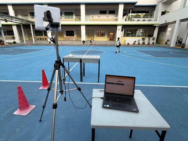 A tripod-mounted RFID reader connected by USB to a teacher’s laptop displaying Mangostin in a school parade square