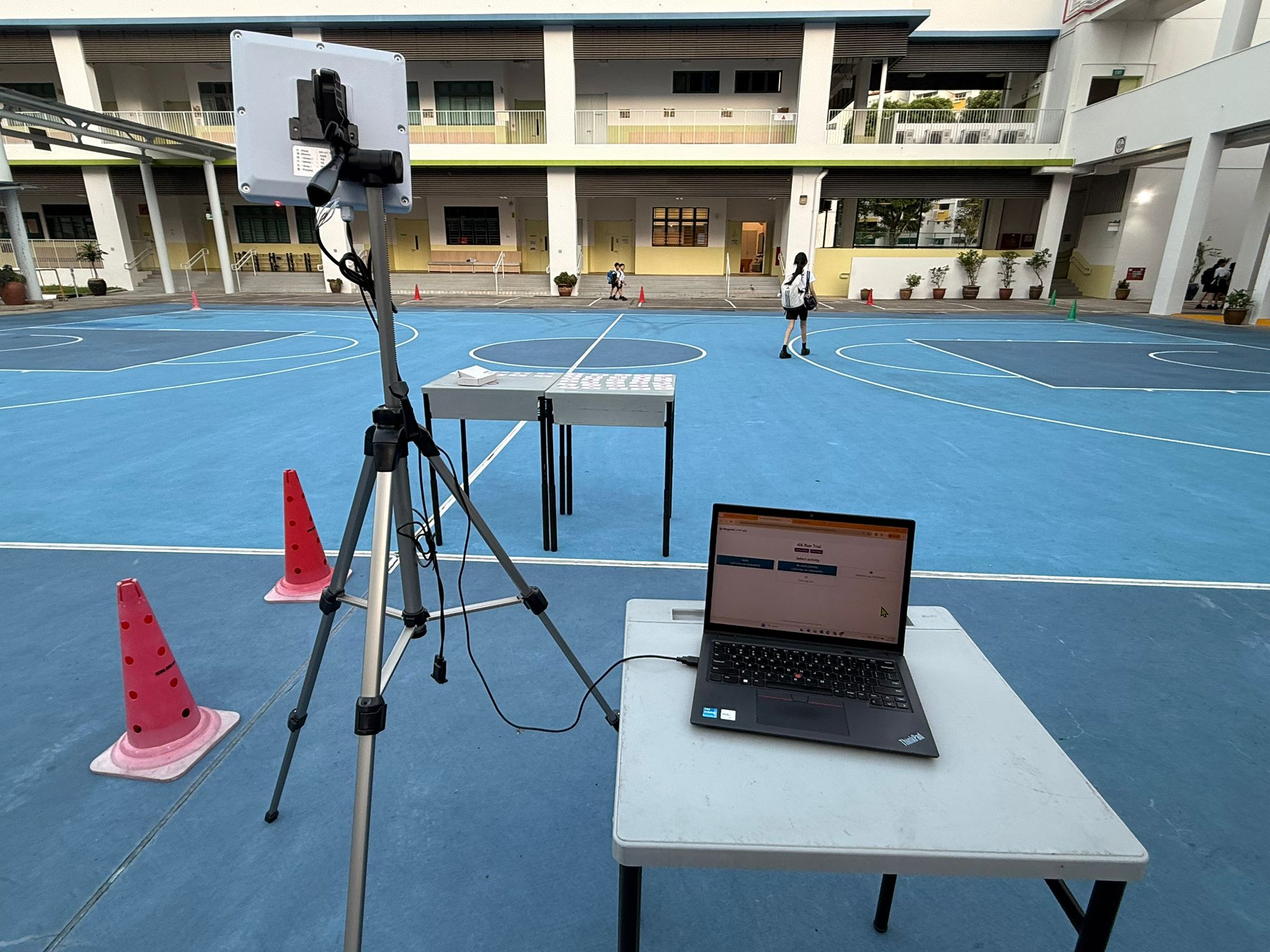 A tripod-mounted RFID reader connected by USB to a teacher’s laptop displaying Mangostin in a school parade square